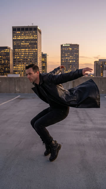 Man in a long black coat in a dodging pose on a city rooftop at dusk